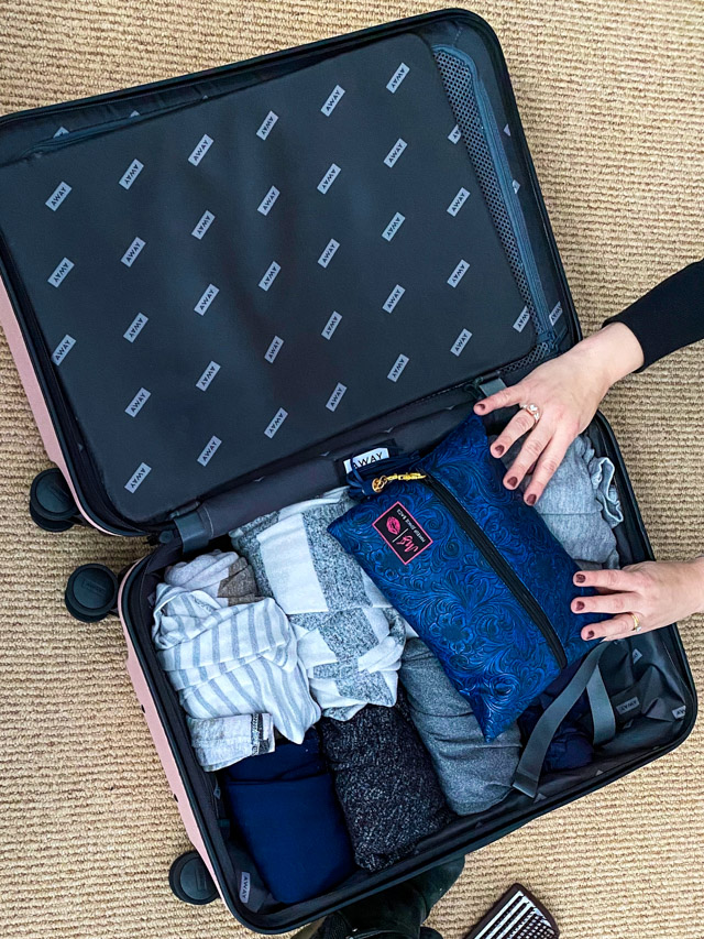 Open suitcase on the floor with hands putting toiletry bag inside.