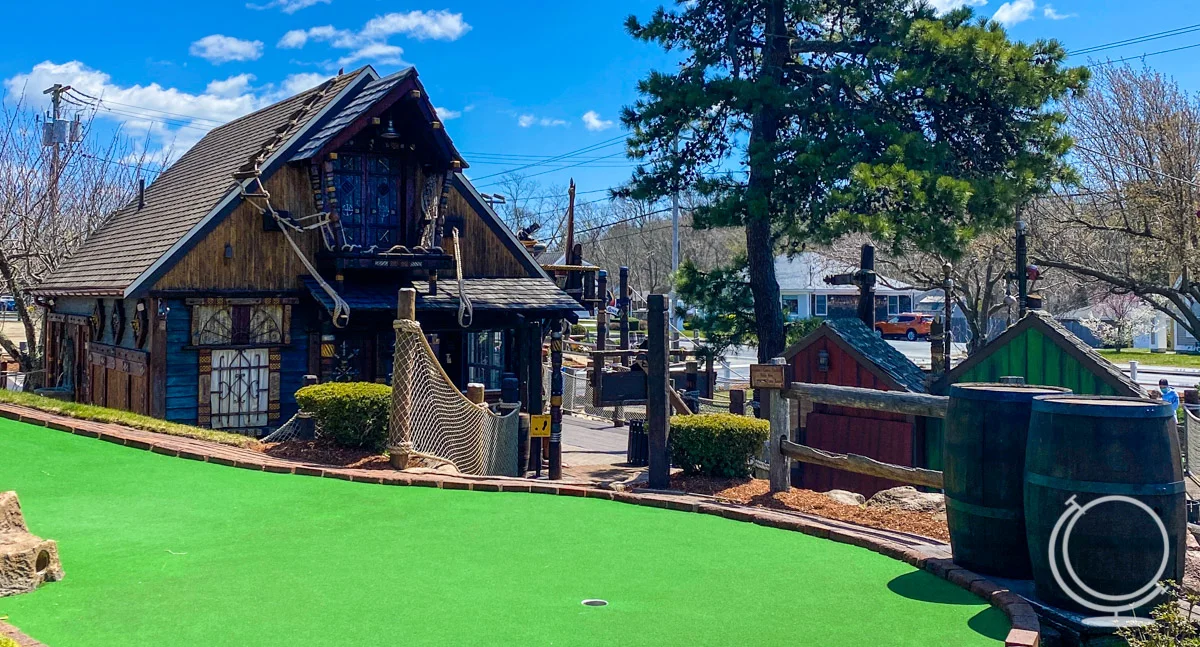 Mini Golf hole in front of several buildings/structures a Pirate's Cove Adventure Golf, the best mini golf on Cape Cod