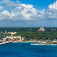 Overview of the Costa Maya cruise terminal from a ship. It shows palm trees, light buildings, straw huts, and a Mayan pyramid in the distance.