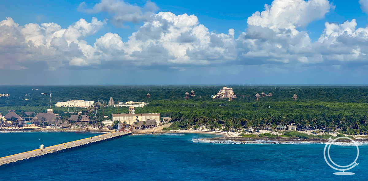 Overview of the Costa Maya cruise terminal from a ship. It shows palm trees, light buildings, straw huts, and a Mayan pyramid in the distance. 