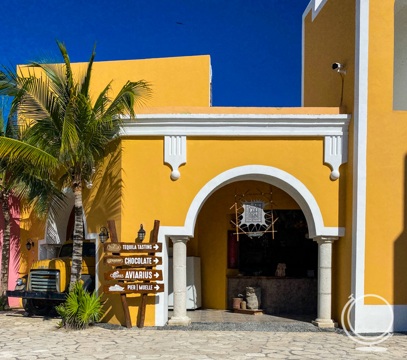 A bright yellow building with a sign showing tequila tasting, chocolate, aviarius, and pier. 