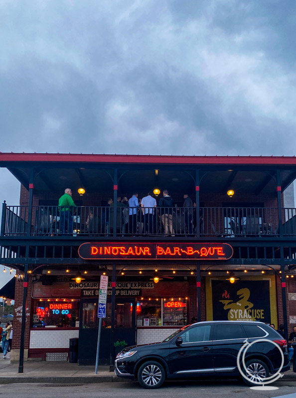 Dinosaur BBQ with neon lights, people on the top balcony, and car in front. 