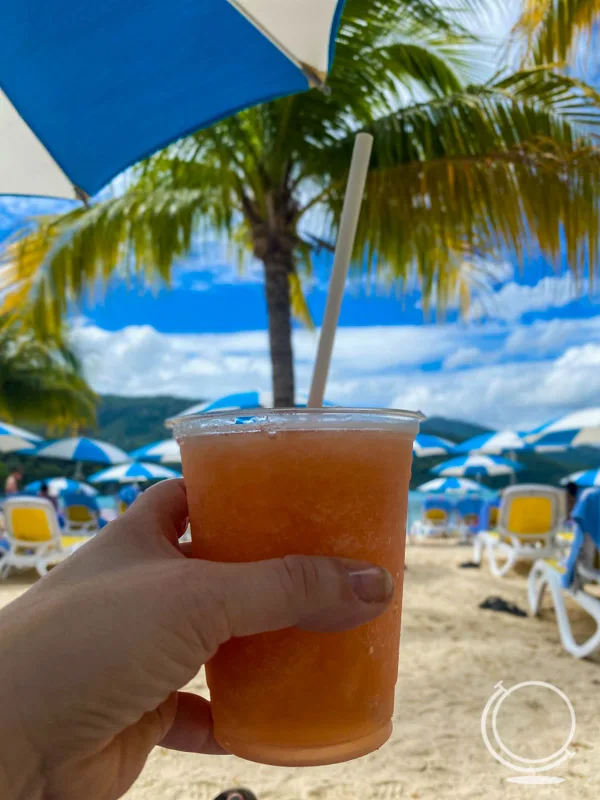 Pink frozen Labadoozie drink held in front of a beach with chairs, umbrellas, and palm trees. 