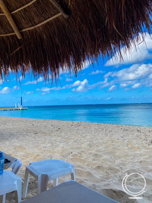 Beach with clear blue water, a small pier, a hint of a natural umbrella showing, and a plastic table. 