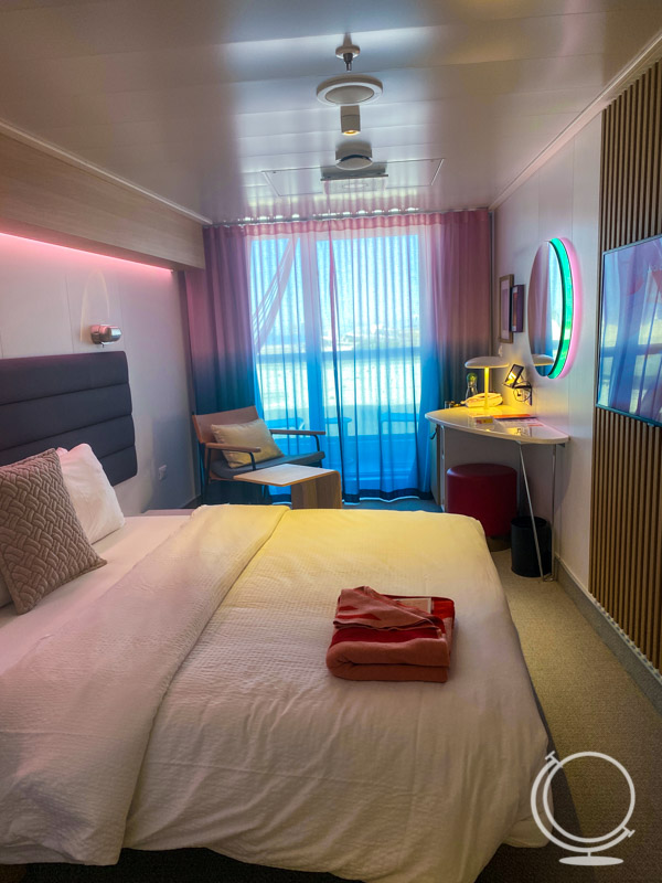 Cabin on the Scarlet Lady, with king bed, small desk, chair, and table as well as a balcony. 