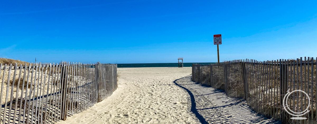 Empty sandy beach with wooden fencing on either side of a sandy path leading to the water. 