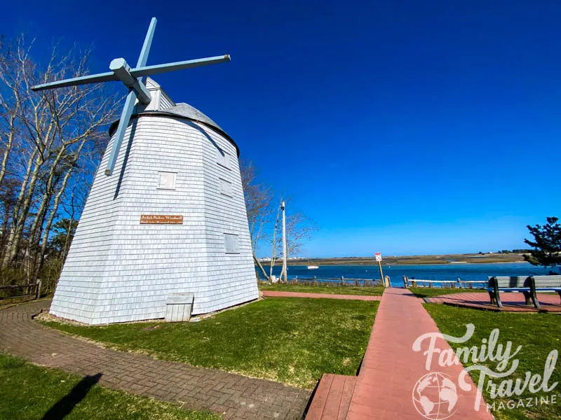 Gray windmill in front of small beach with brick paths and wooden bench