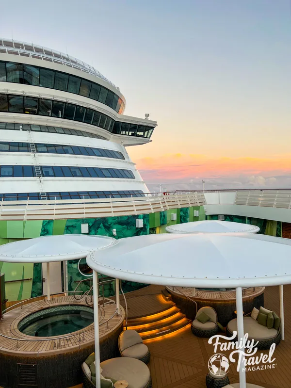 Rainforest room outdoor area with hot tubs and sun loungers with a pink sky outside along the front of the ship. 