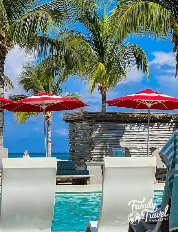 Beach Club at Bimini with red and white umbrellas, palm trees, and pool chairs in front of a chair. 
