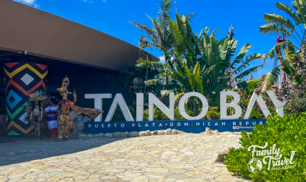 Large sign advertising Taino Bay cruise port with people in local costumes and greenery around it. 