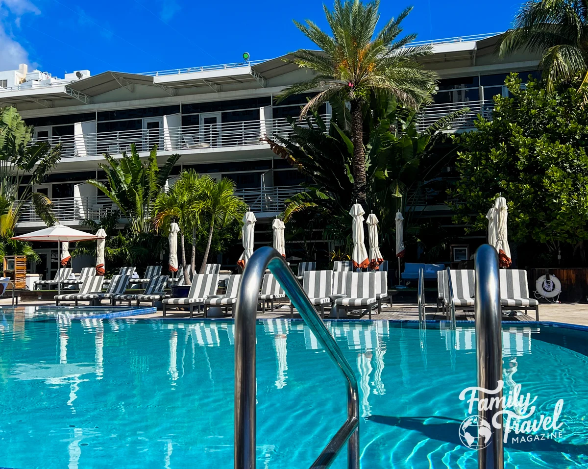 Pool surrounded by palm trees, chairs, and umbrellas, in front of the hotel 