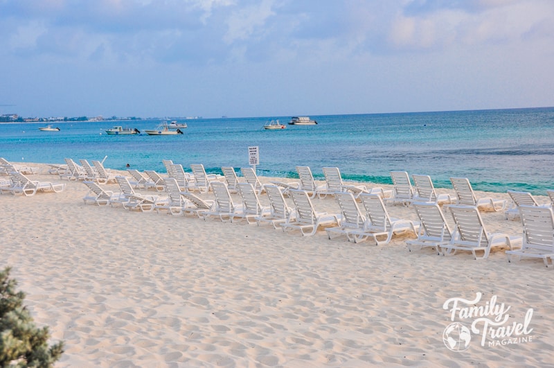empty beach chairs in the sand in front of turquoise water with boats in it
