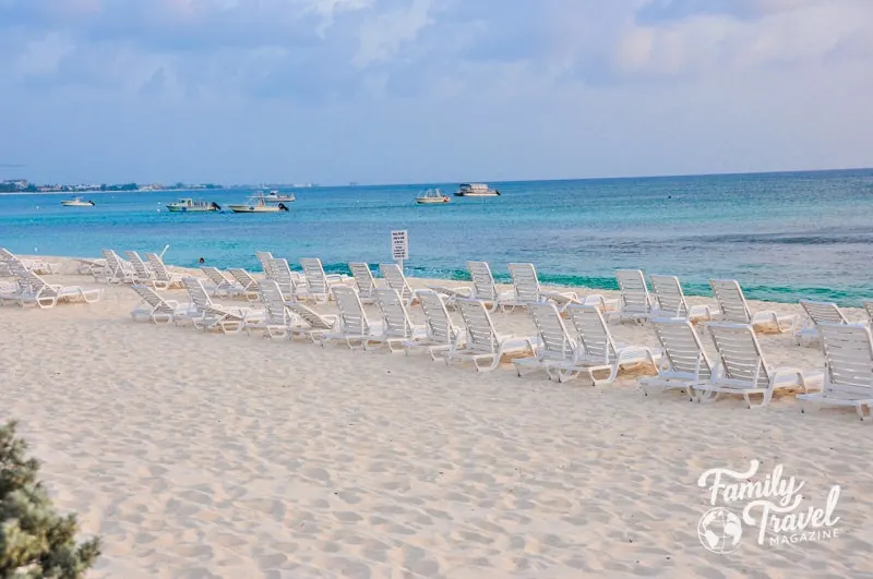 empty beach chairs in the sand in front of turquoise water with boats in it