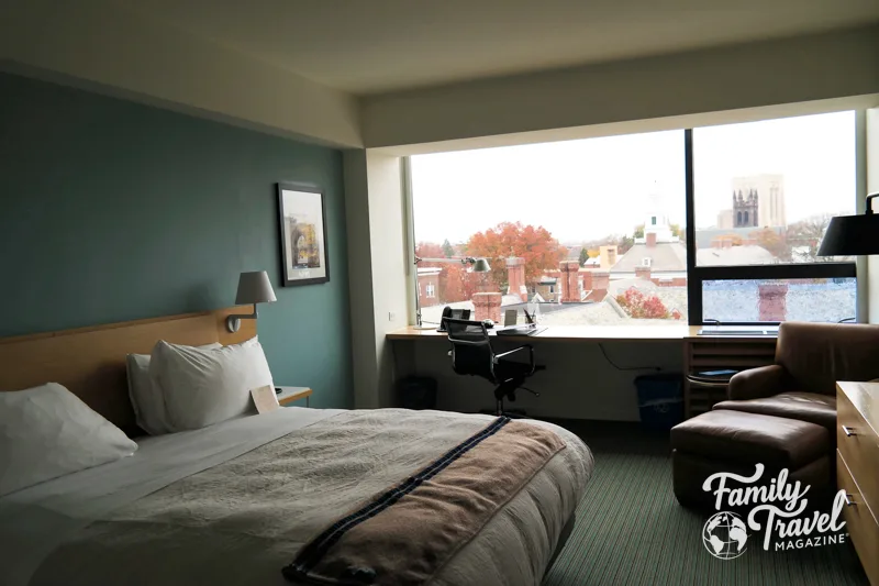 buildings and fall foliage as seen through hotel window with desk, dresser, chair, and bed. 