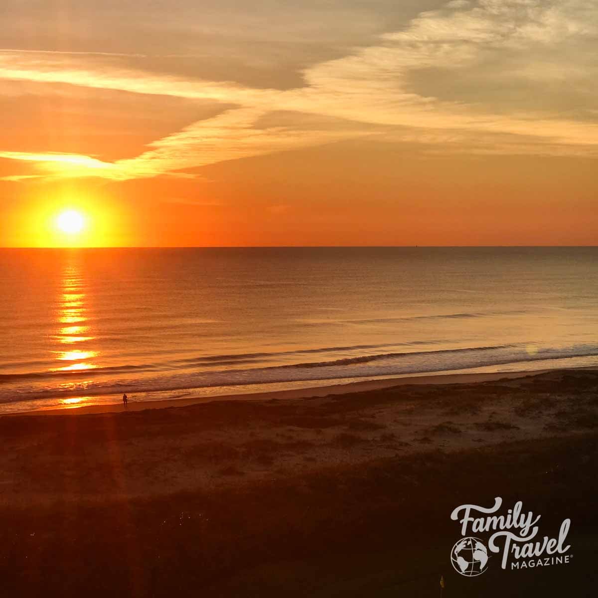 Bright orange sunrise over a calm ocean beach with a person walking on the sand.