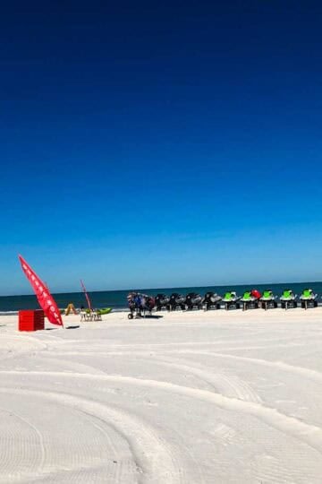 White sand beach at Clearwater Beach, Florida, with Pier 60, jet skis, and a rental booth under a clear blue sky.