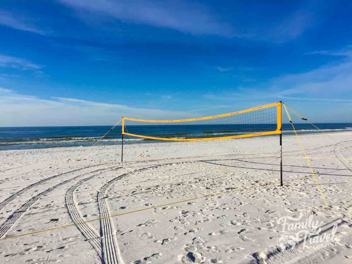 A yellow volleyball net stands on a white sand beach with ocean waves under a clear blue sky.
