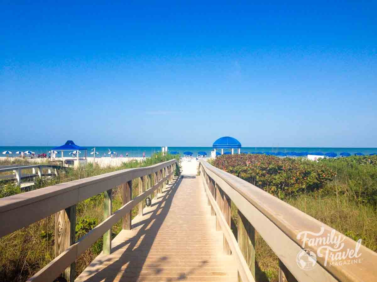 Wooden boardwalk leads to Marco Island Beach featuring blue umbrellas and a gazebo.