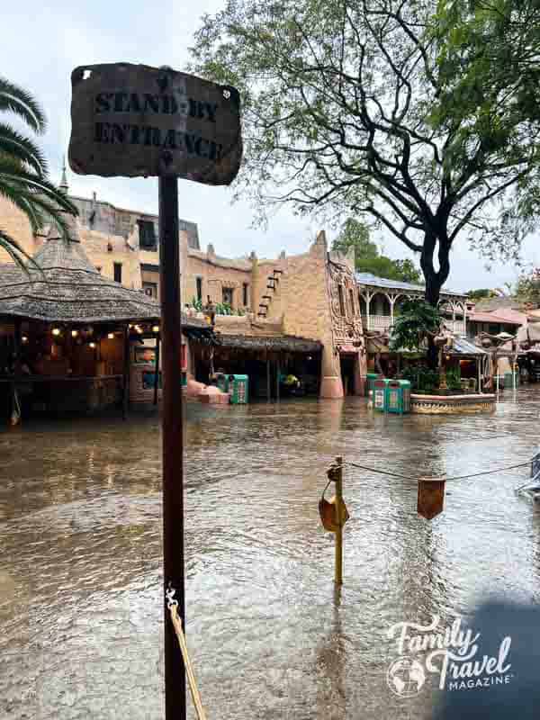 Rainy day in Adventureland at Disneyland, with puddles covering the ground near the standby entrance and tropical-themed buildings in the background.