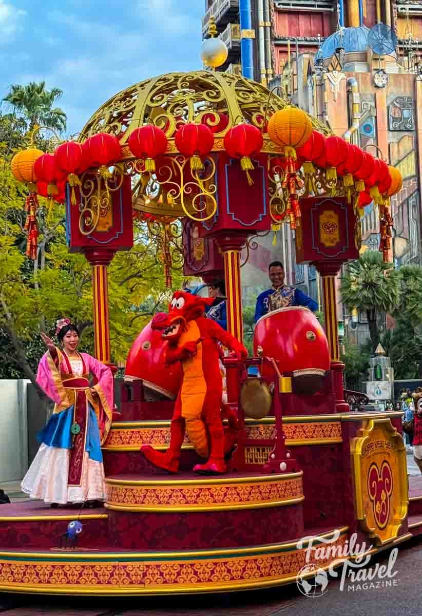 Mulan and Mushu perform on a vibrant Lunar New Year parade float at Disney California Adventure, decorated with red lanterns, gold details, and giant drums.