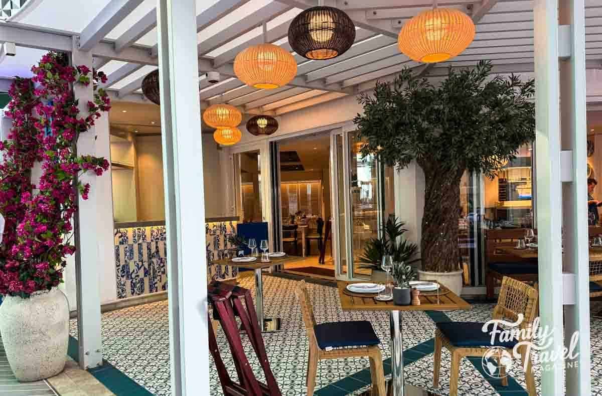 Outdoor seating area of a Mediterranean-style restaurant with wooden tables set for two, surrounded by potted plants, pink bougainvillea, and hanging wicker lanterns. The space features blue and white tile accents, patterned flooring, and a tree in a large planter, creating a cozy and elegant ambiance.
