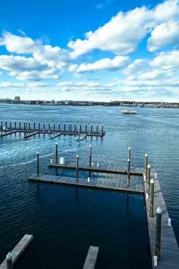 A scenic waterfront view from the Institute of Contemporary Art in Boston, showing a marina with boats, empty docks, and the expansive blue waters of Boston Harbor under a partly cloudy sky. The American flag waves on the left, and a walkway along the rocky shoreline is visible on the right. The image is branded with the Family Travel Magazine logo in the bottom right corner.