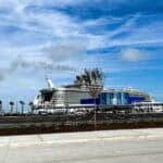 A large Royal Caribbean cruise ship docked at the Galveston cruise terminal under a bright blue sky, with a nearly empty parking lot and newly planted trees in the foreground. The terminal building features Royal Caribbean branding, and light smoke trails can be seen from the ship's funnel.