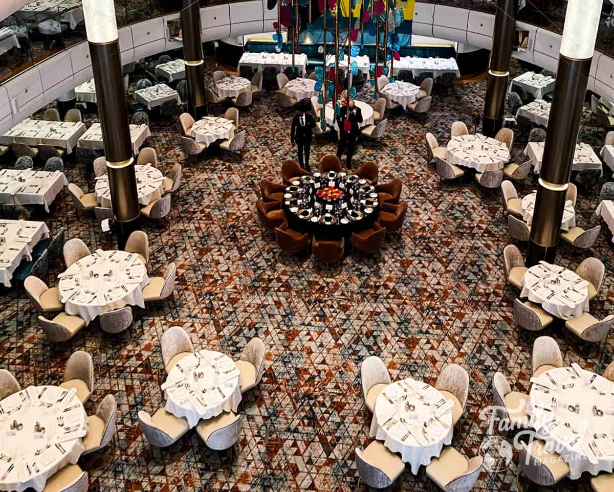 Overhead view of the main dining room on a Royal Caribbean cruise ship, featuring multiple round tables elegantly set with white tablecloths and silverware. The room has a patterned carpet, modern pillars, and a colorful hanging sculpture in the center. A large circular table surrounded by brown chairs sits in the middle, with servers preparing for service. The “Family Travel Magazine” watermark appears in the bottom right corner.