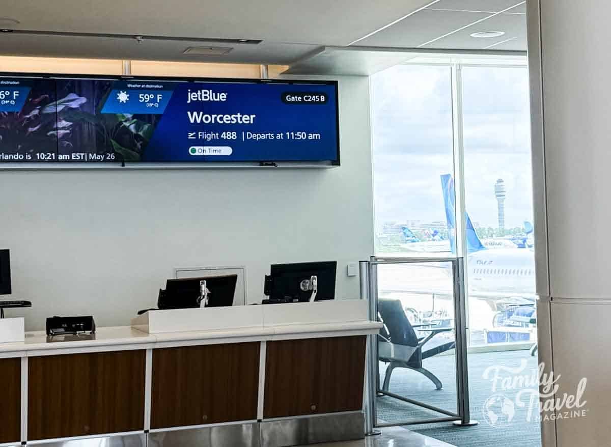 JetBlue departure gate at Orlando International Airport (MCO) for Flight 488 to Worcester, Massachusetts. Digital gate sign displays flight details and weather, with JetBlue aircraft visible through the terminal window. Family Travel Magazine watermark in corner.