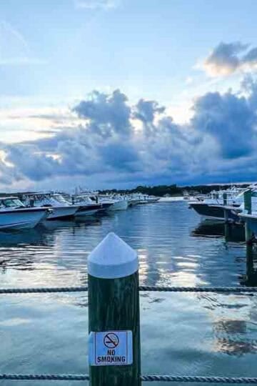 Scenic view of a marina at dusk with rows of docked motorboats on calm water, under a dramatic cloudy sky. A wooden post in the foreground displays a “No Smoking” sign, and the Family Travel Magazine logo is in the bottom right corner.