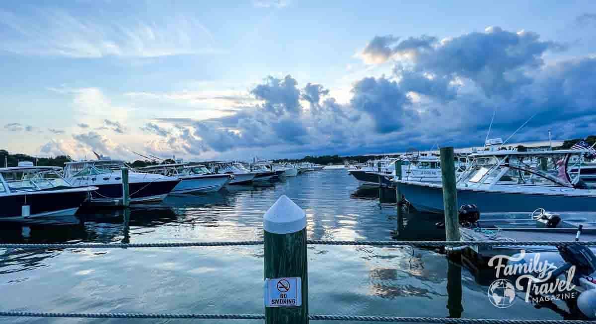 Scenic view of a marina at dusk with rows of docked motorboats on calm water, under a dramatic cloudy sky. A wooden post in the foreground displays a “No Smoking” sign, and the Family Travel Magazine logo is in the bottom right corner.