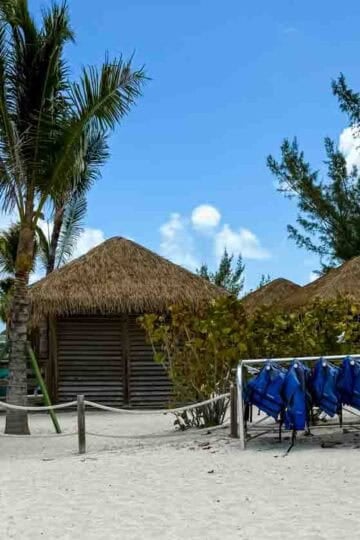 Cabana check-in area on a tropical beach at a private cruise line island, featuring thatched-roof cabanas, palm trees, turquoise cabana check-in booth, and racks of red and blue life jackets under a sunny blue sky.