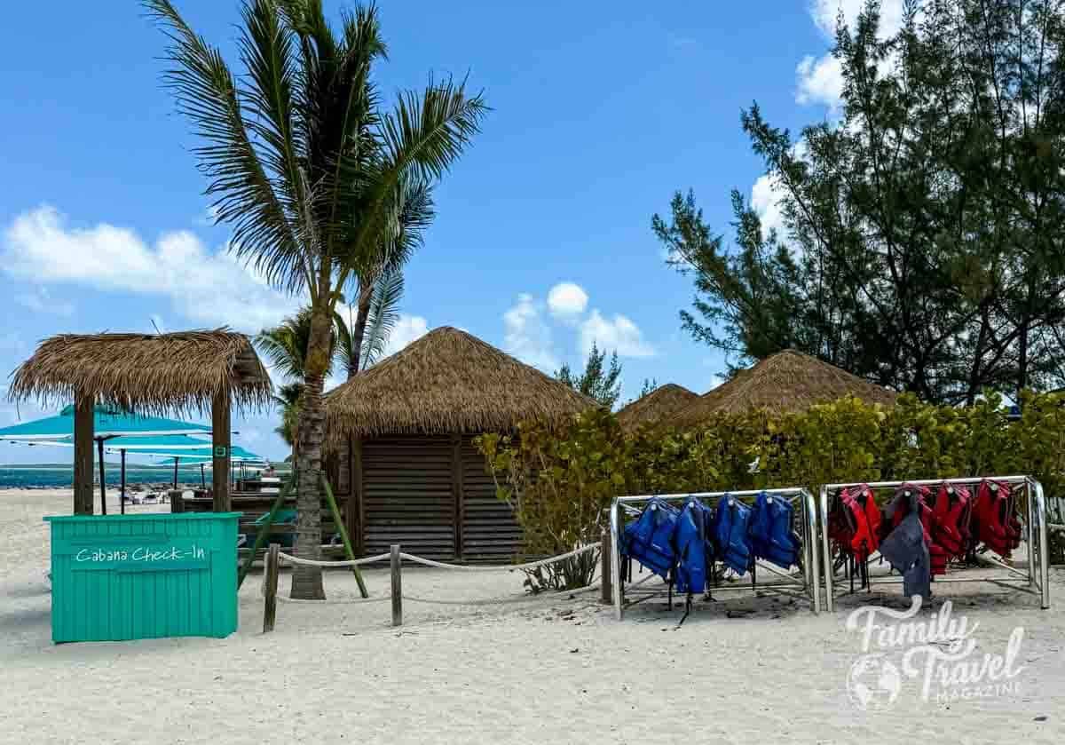 Cabana check-in area on a tropical beach at a private cruise line island, featuring thatched-roof cabanas, palm trees, turquoise cabana check-in booth, and racks of red and blue life jackets under a sunny blue sky.