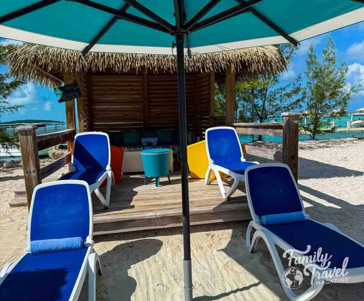 Private beach cabana with thatched roof at a cruise line’s private island, featuring blue lounge chairs, colorful seating, and a large umbrella on white sand overlooking turquoise ocean water