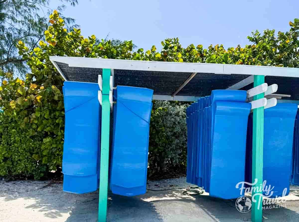 Blue floating mats stored under a shaded rack at a cruise line’s private island beach, with lush greenery and clear blue sky in the background.