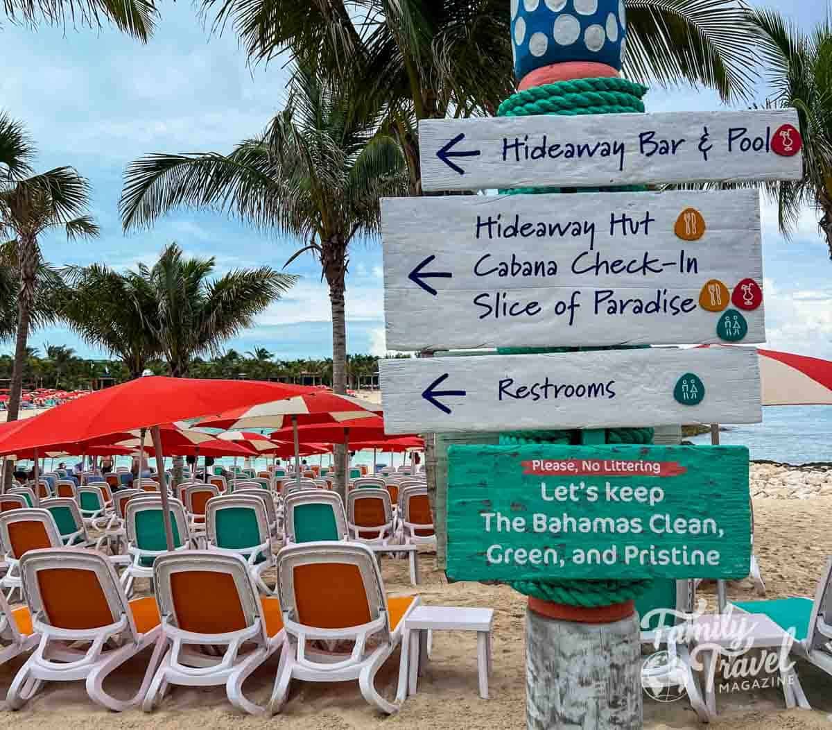 Beach signpost at a cruise line’s private island in the Bahamas, pointing to Hideaway Bar & Pool, cabana check-in, and restrooms, with palm trees, red umbrellas, and rows of lounge chairs on sandy beach in the background.
