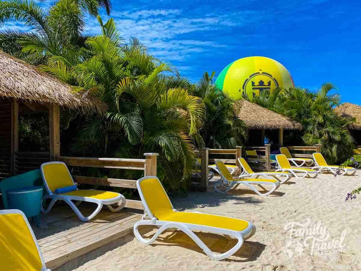 Private cabanas and yellow lounge chairs on a sandy beach at a cruise line’s private island, with lush palm trees and the signature Royal Caribbean helium balloon in the background under a bright blue sky.