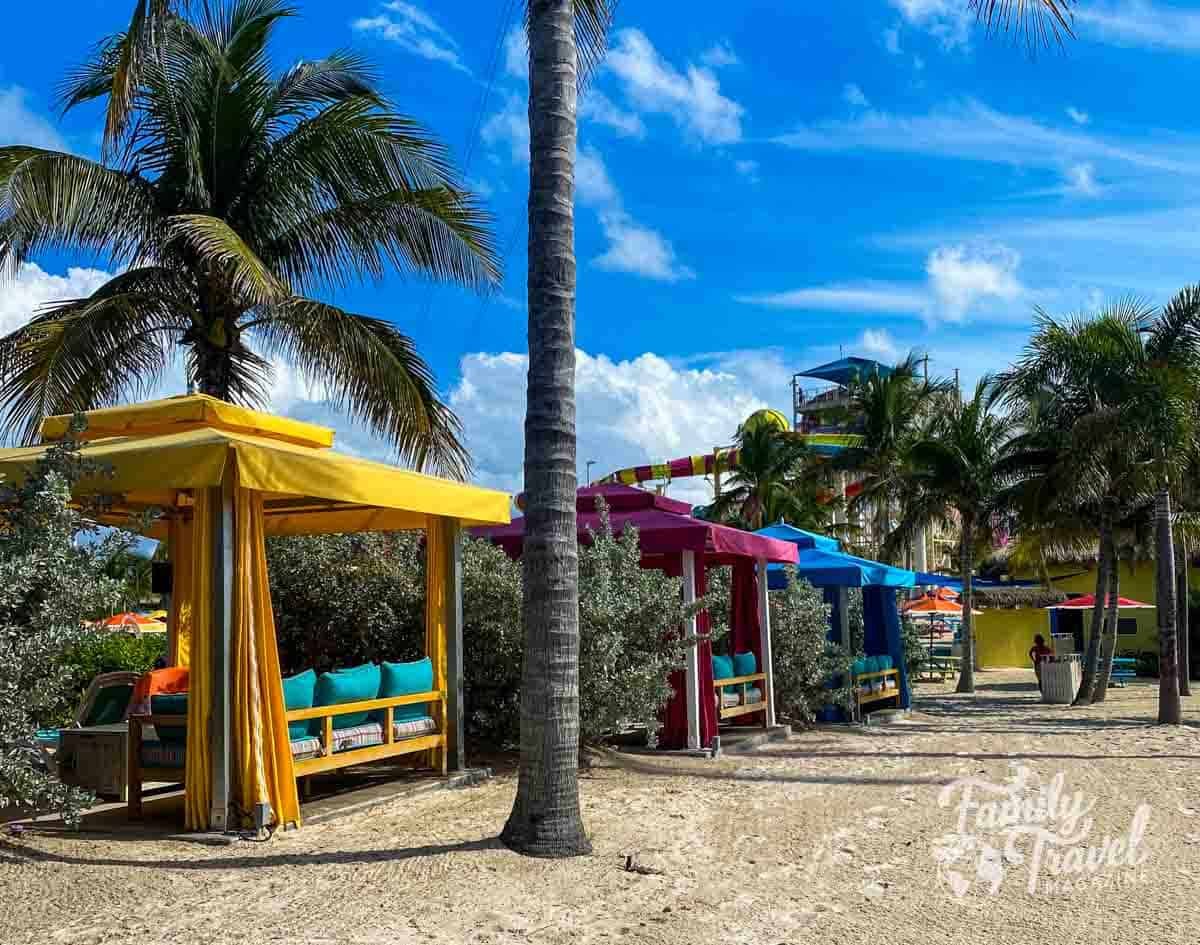 Colorful private cabanas in yellow, red, and blue at a water park on a cruise line’s private island, with palm trees, sandy pathways, and waterslides visible in the background under a bright blue sky.