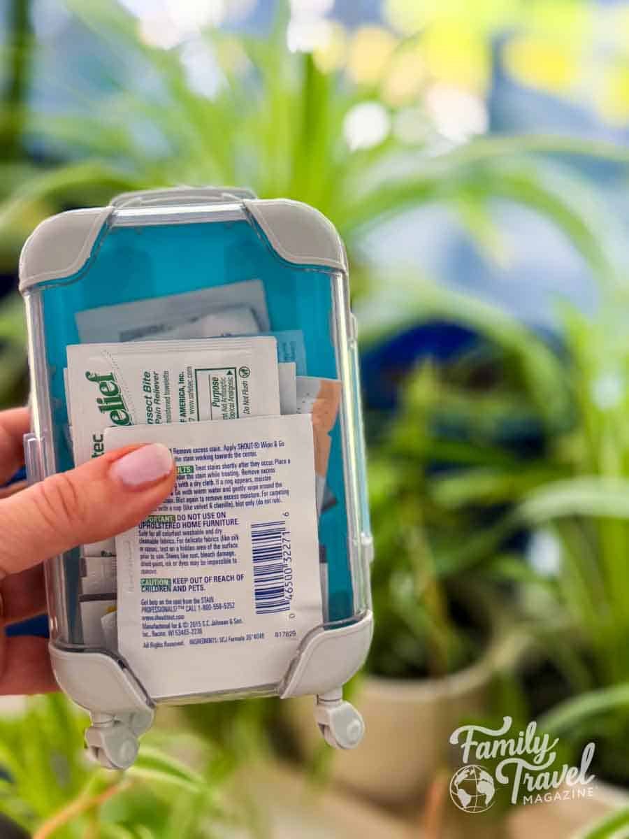 Hand holding a mini suitcase-shaped travel essentials kit filled with wipes and first aid supplies, with green plants in the background—perfect for family travel preparedness