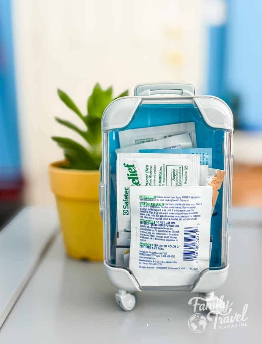 Compact travel essentials kit shaped like a mini suitcase, filled with wipes and first aid items, standing on a table with a yellow potted succulent in the background—ideal for organized family travel.