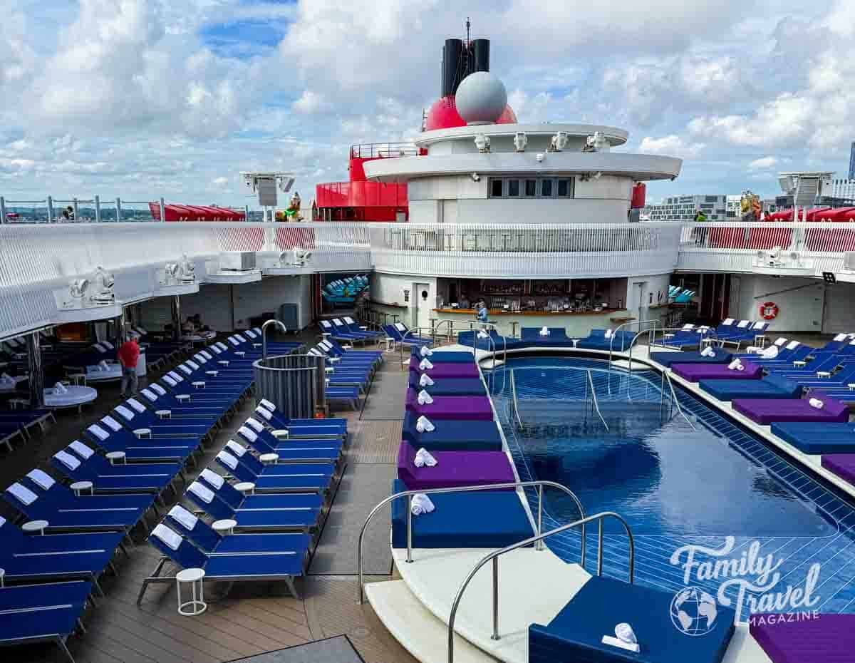 Athletic Club pool deck on Virgin Voyages’ Brilliant Lady with blue and purple lounge chairs, swimming pool, and poolside bar under a partly cloudy sky.