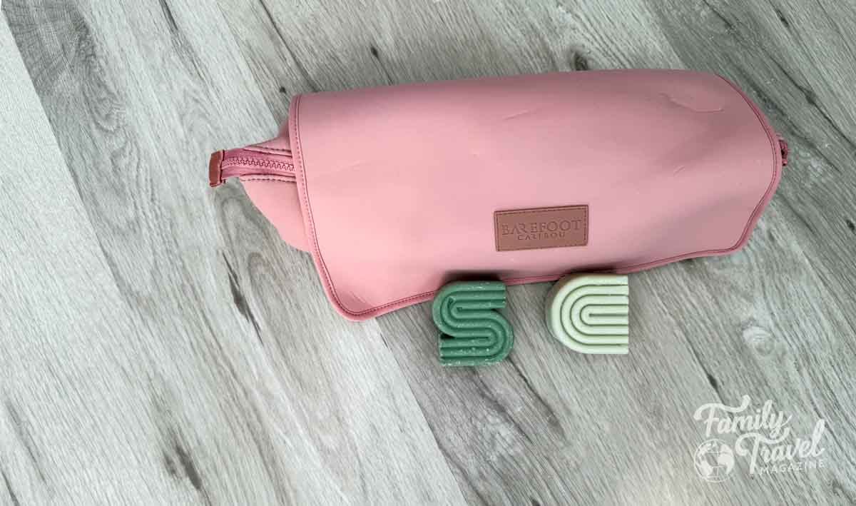 Eco-friendly shampoo and conditioner bars in green and light gray placed in front of a pink hair product bag on a wood floor.