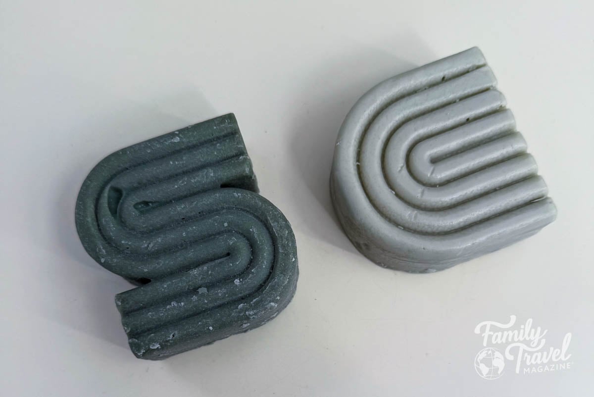 Solid shampoo and conditioner bars in dark green and light gray, shaped with modern curved grooves, sitting on a white surface.