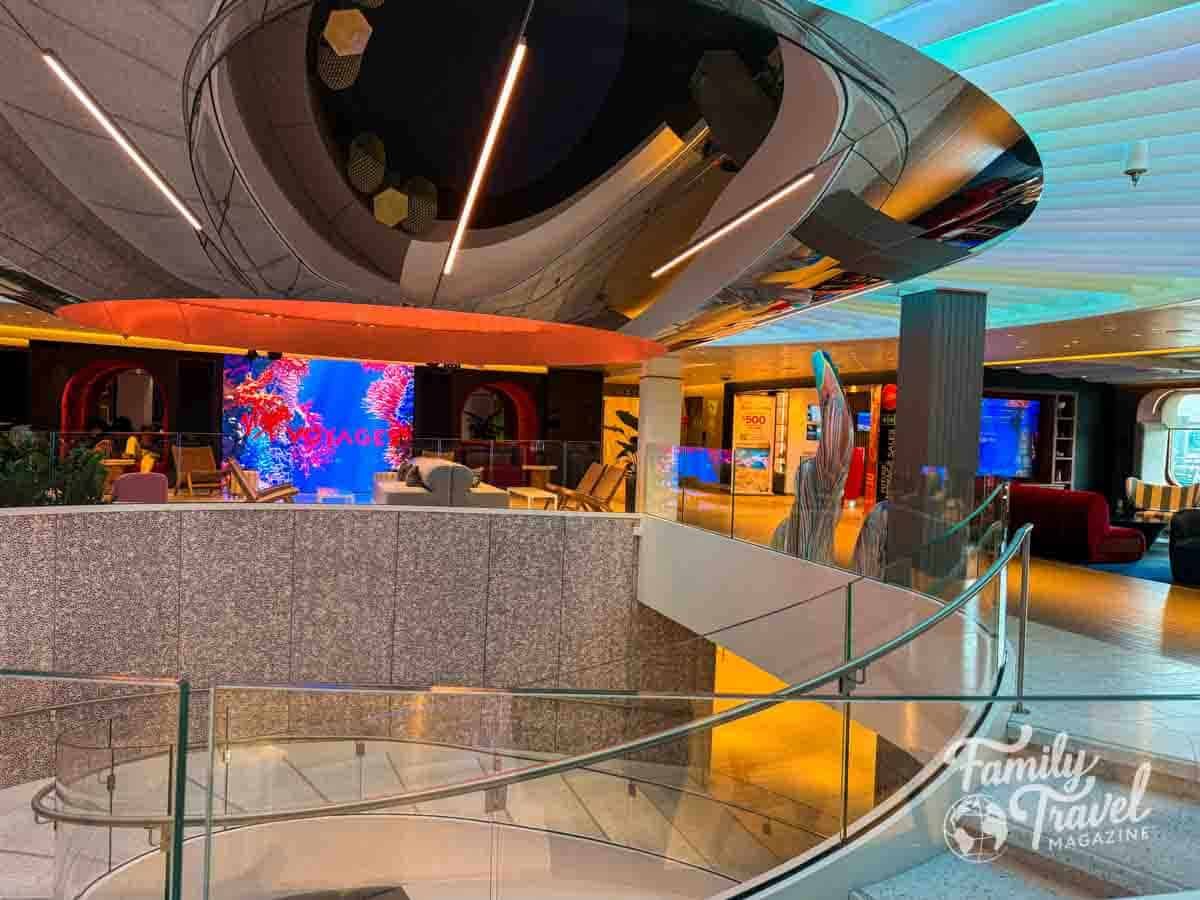 The Roundabout atrium on Virgin Voyages’ Brilliant Lady cruise ship with modern design, curved staircase, digital screens, and lounge seating.