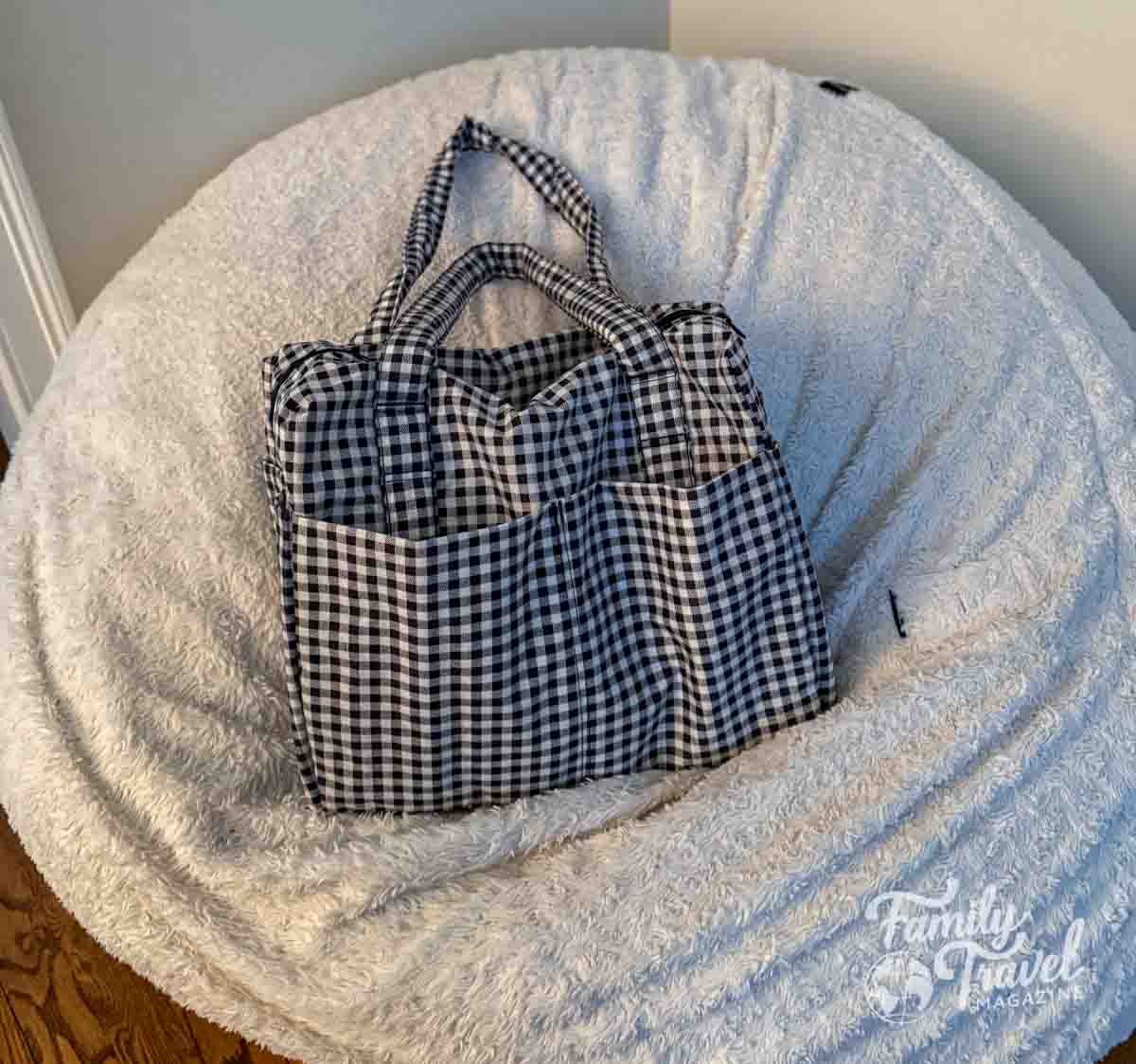 A black and white gingham Baggu Cloud tote bag sits on a white sherpa bean bag chair, showing a lightweight and casual option for travel or everyday use.