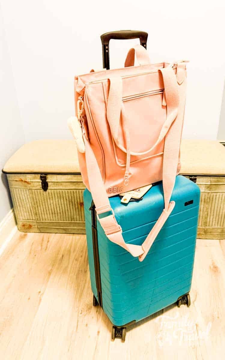 A blush pink Beis North South tote bag sits on top of a teal carry-on suitcase with a telescoping handle, ready for travel, in a bright entryway with a wooden floor.