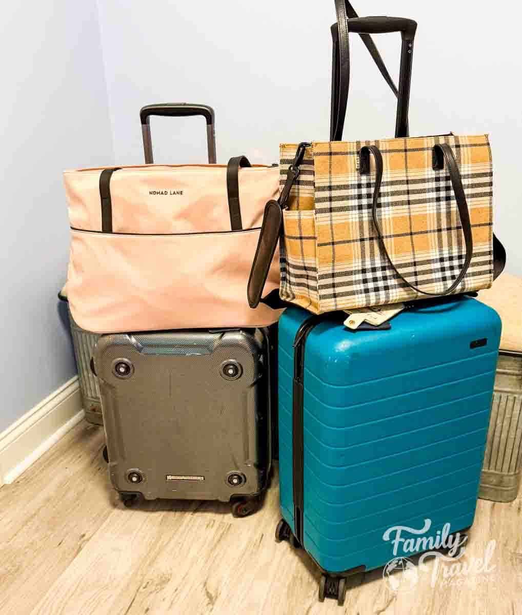 Two stylish travel totes, a blush pink Nomad Lane bag and a tan plaid Beis tote, rest on top of a teal suitcase, perfect for organized carry-on travel.