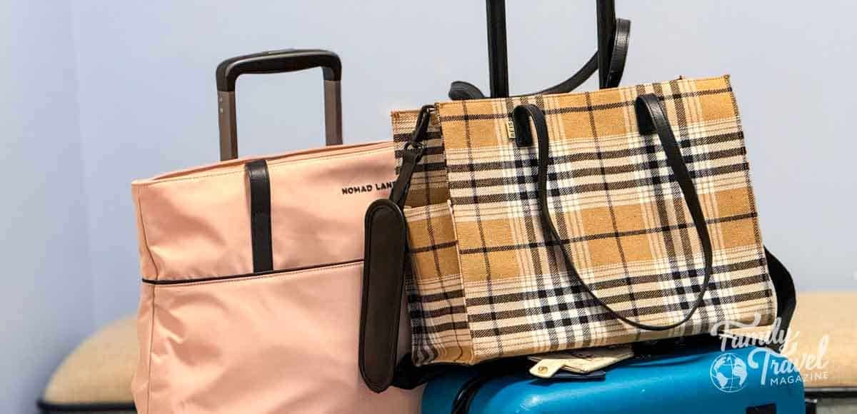 Two stylish travel totes, a blush pink Nomad Lane bag and a tan plaid Beis tote, rest on top of a teal suitcase, perfect for organized carry-on travel.
