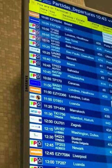 Airport departures board showing flights from Lisbon with destinations including Bristol, Dublin, London Heathrow, Newark, Miami, Boston, and more, displayed on a digital screen at 10:43 a.m. with various flight statuses in Portuguese. The Family Travel Magazine logo appears at the bottom right corner.