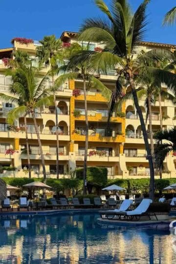 View of Velas Vallarta resort with palm trees, colorful flowers on balconies, and a large pool reflecting the yellow building under a bright blue sky.